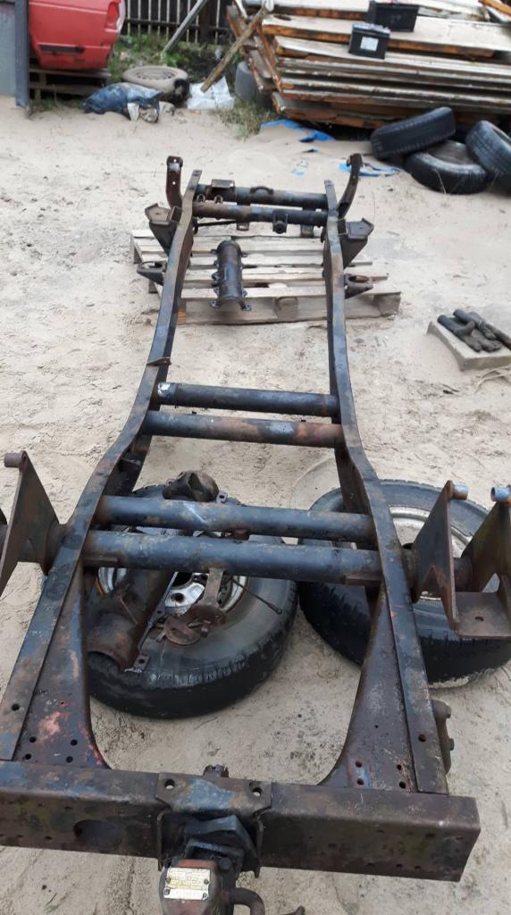 Metalowa rama samochodu z widocznymi śladami korozji i spawania, umieszczona na drewnianej palecie na piaszczystym podłożu, z oponami w tle.