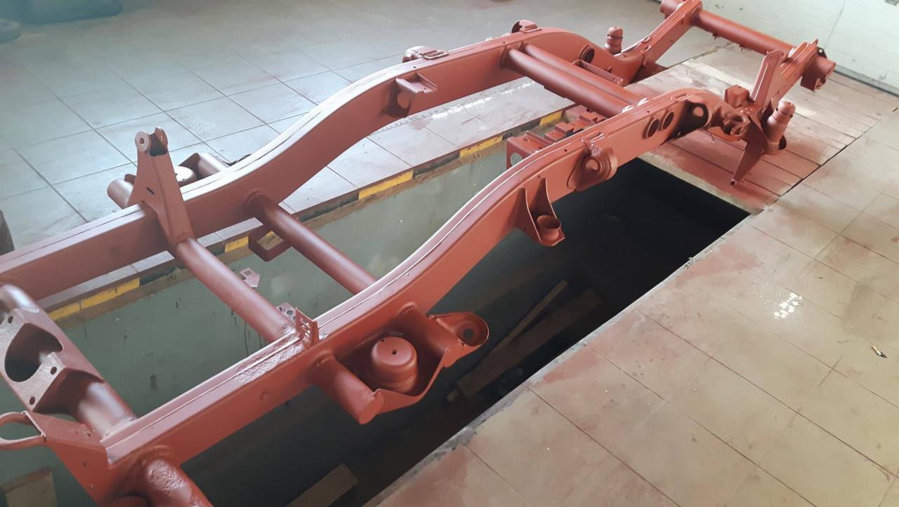 Czerwona, pomalowana rama samochodu leży na podłodze warsztatu nad kanałem. Widoczne ślady farby na podłodze.