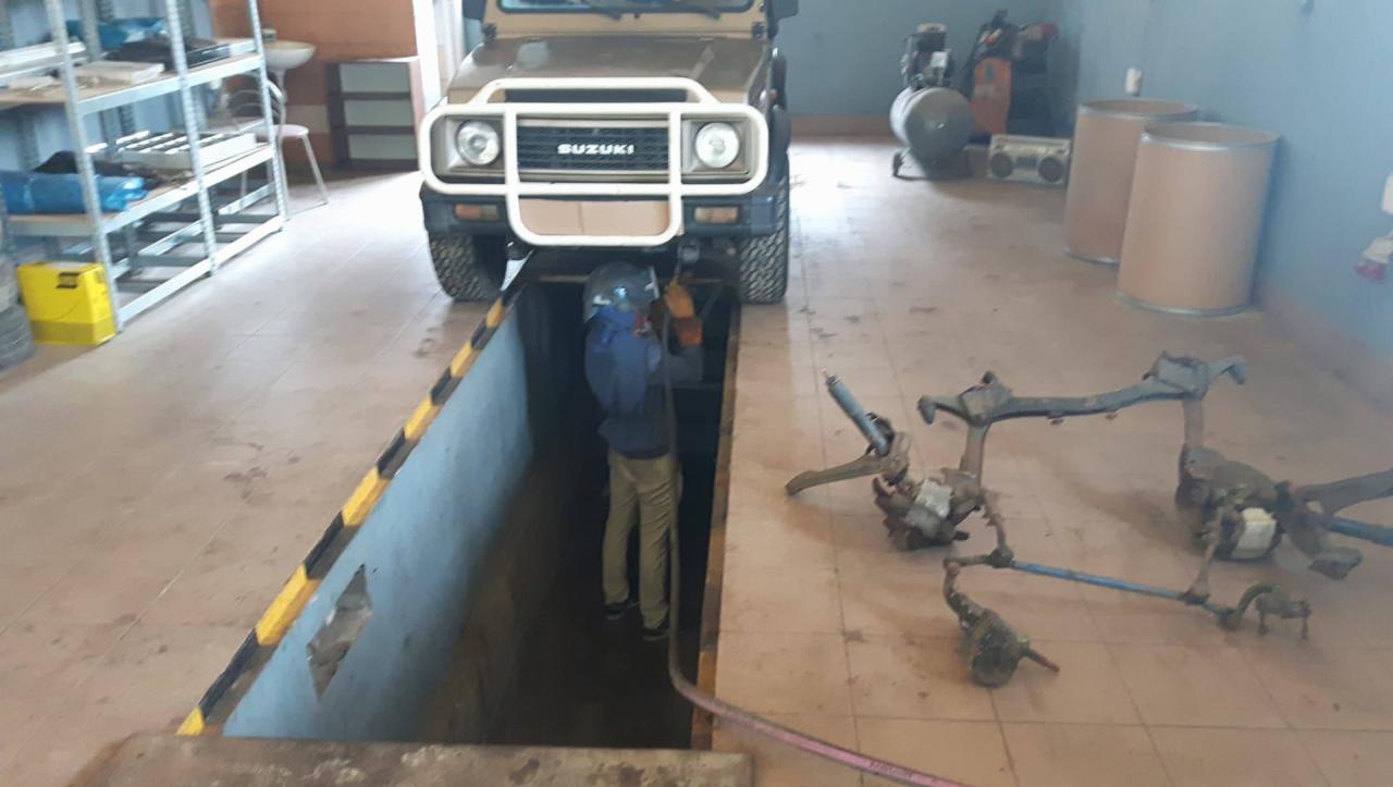 Mechanik w kanale naprawczym pod samochodem Suzuki, spawanie podwozia, w tle części samochodowe i narzędzia w warsztacie.