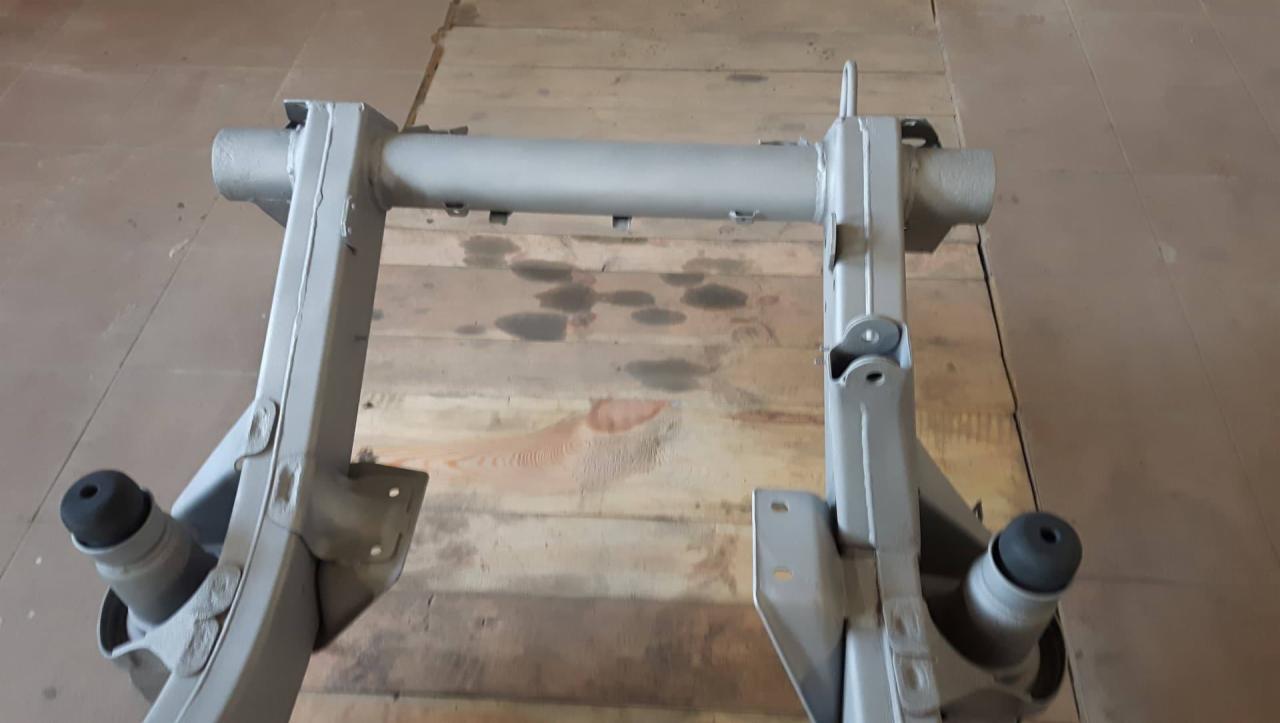 Metalowa rama konstrukcyjna, prawdopodobnie podwozia pojazdu, pomalowana na szaro, leżąca na drewnianej podłodze. Widoczne spawy i mocowania.