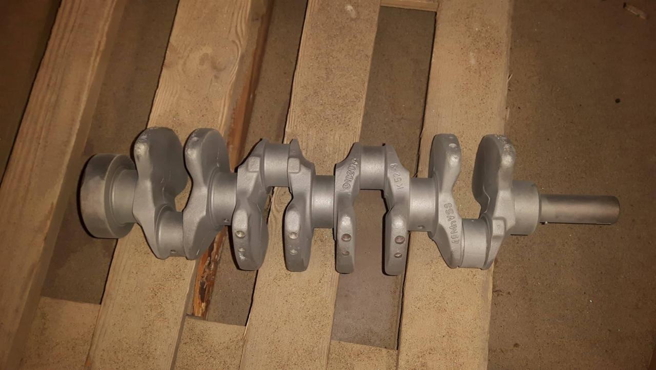 Szary, surowy wał korbowy leżący na drewnianej palecie, widoczne oznaczenia 49MnVS3, K52-8 oraz 010201.