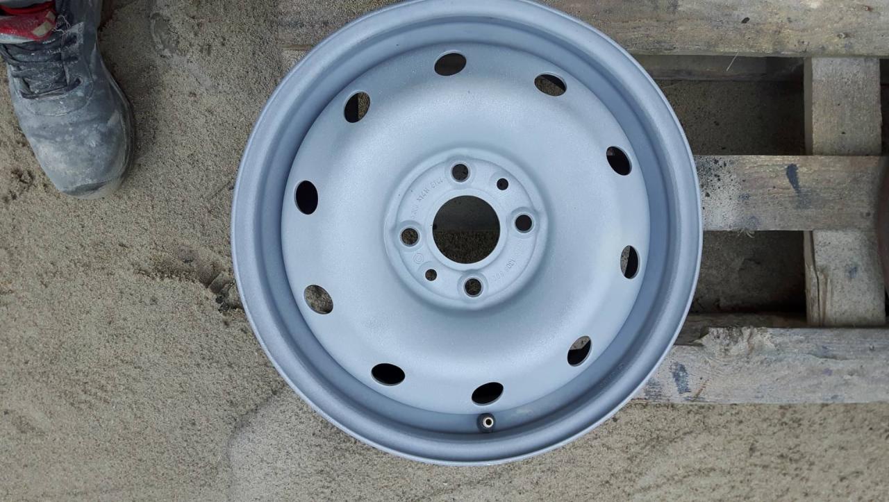 Pomalowana na szaro felga samochodowa leży na piasku obok roboczego buta i drewnianej palety.