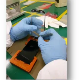 Naprawa ekranu telefonu komórkowego w sterylnych warunkach, osoba w rękawicach i kombinezonie ochronnym precyzyjnie nakłada nową szybkę za pomocą pęsety.