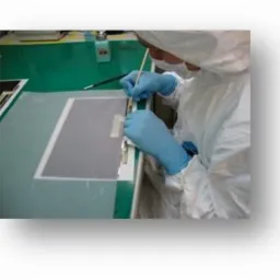 Pracownik w kombinezonie ochronnym i niebieskich rękawicach precyzyjnie nakleja etykietę na płaski, szary element na stole w sterylnym pomieszczeniu.