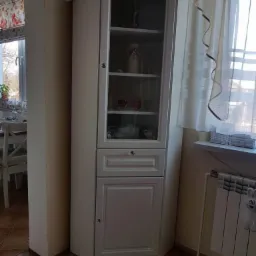 Biała, narożna witryna z przeszkloną górną częścią i dolną szafką, uzupełniona szufladą, stoi w rogu pokoju na parkiecie, obok grzejnika i okna z firanką.
