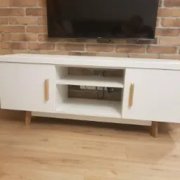 Biała szafka RTV z drewnianymi uchwytami i nóżkami, stojąca na drewnianej podłodze przed ceglaną ścianą, z widocznym telewizorem na ścianie.