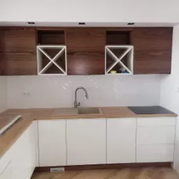 Nowoczesna zabudowa kuchenna z białymi, lakierowanymi frontami szafek dolnych i drewnianymi szafkami górnymi z otwartymi półkami na wino, jasny blat drewniany, białe płytki ułożone w jodełkę...