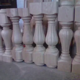 Surowe, drewniane balustrady schodowe o różnych kształtach, ustawione w rzędzie na szarym tle, przed obróbką wykończeniową.