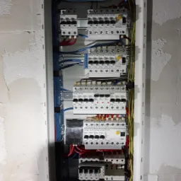 Rozdzielnica elektryczna z widocznymi bezpiecznikami, przewodami o różnych kolorach i listwą zaciskową, zamontowana na surowej ścianie w trakcie prac wykończeniowych.