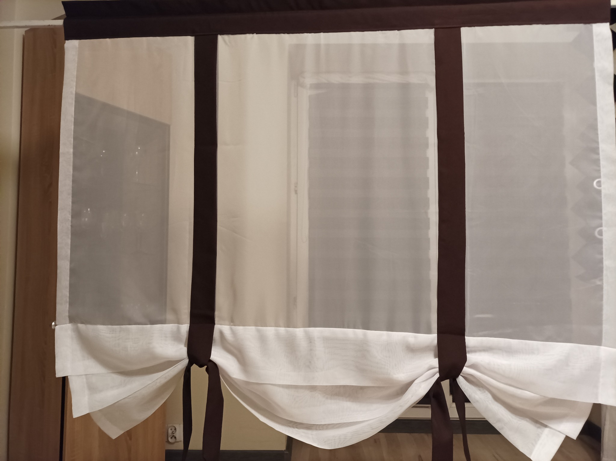 Delikatna, biała, podwieszana roleta rzymska z brązowymi akcentami, zamontowana w oknie.