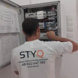 Elektryk w firmowej koszulce STYQ pracuje przy otwartej skrzynce elektrycznej z bezpiecznikami i schematami, widoczne przewody i aparatura modułowa