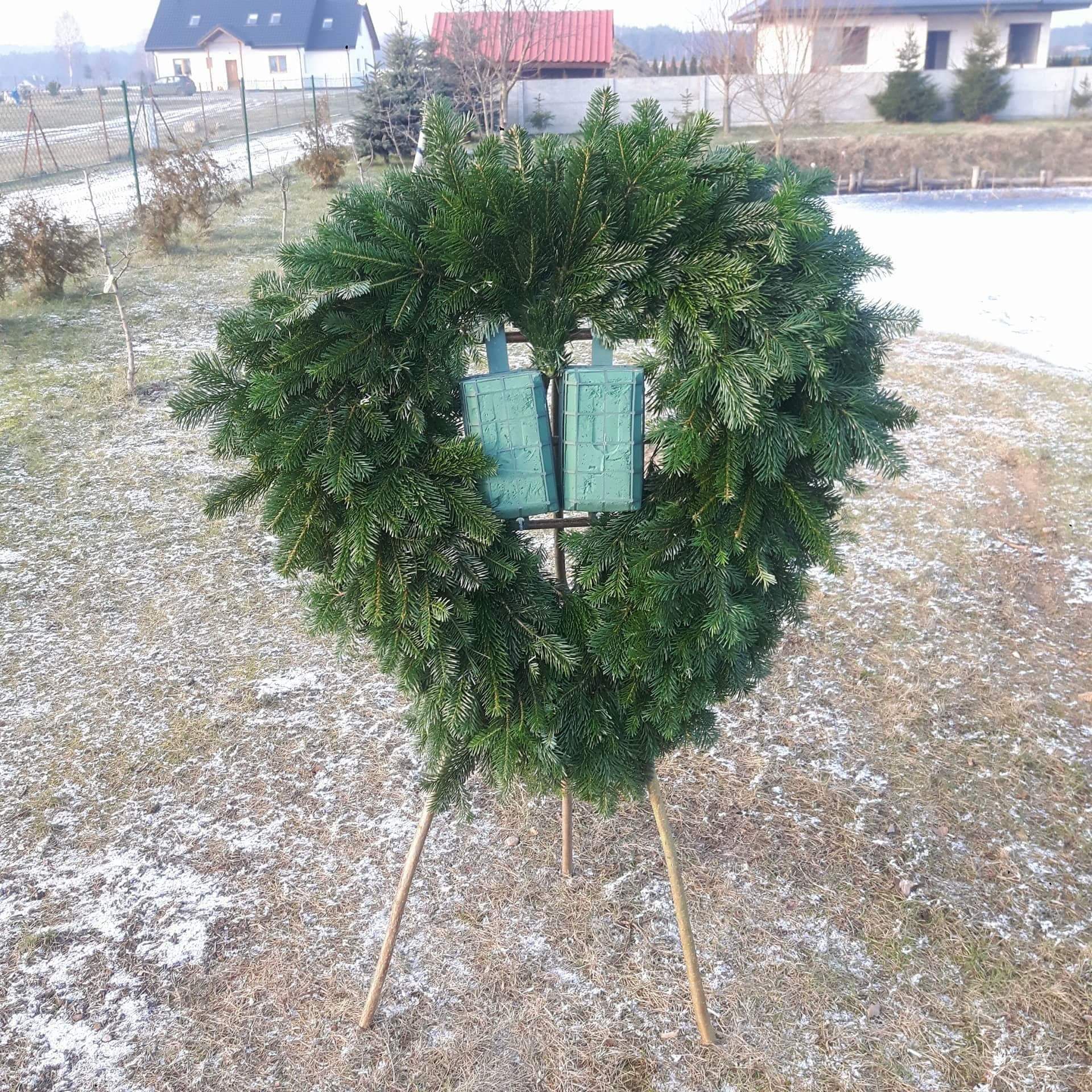 Wieniec z gałązek iglastych na metalowym stelażu z dwoma zielonymi pojemnikami na kwiaty, ustawiony na trawniku z resztkami śniegu, w tle domy jednorodzinne.