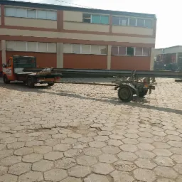 Pomarańczowy samochód dostawczy z przyczepką przewozi długi, stalowy profil na betonowym placu przed budynkiem.