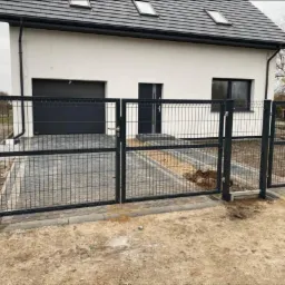 Nowoczesne, grafitowe ogrodzenie z siatki panelowej z bramą i furtką, na tle białego domu z grafitowym dachem i kostką brukową.