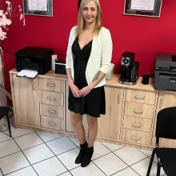 Kobieta w czarnej sukience i białym żakiecie stoi na tle czerwonej ściany z certyfikatami, szafką z drukarkami i ekspresem do kawy.