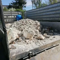 Pełna gruzu skrzynia samochodu ciężarowego, widoczny duży biały fragment betonu i niebieski worek na tle zabudowań i drzew.