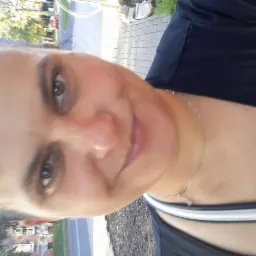 Uśmiechnięta kobieta w sportowym ubraniu, z delikatnym naszyjnikiem, sfotografowana z bliska. Widoczny fragment ulicy i zieleni w tle, zdjęcie obrócone o 90 stopni.