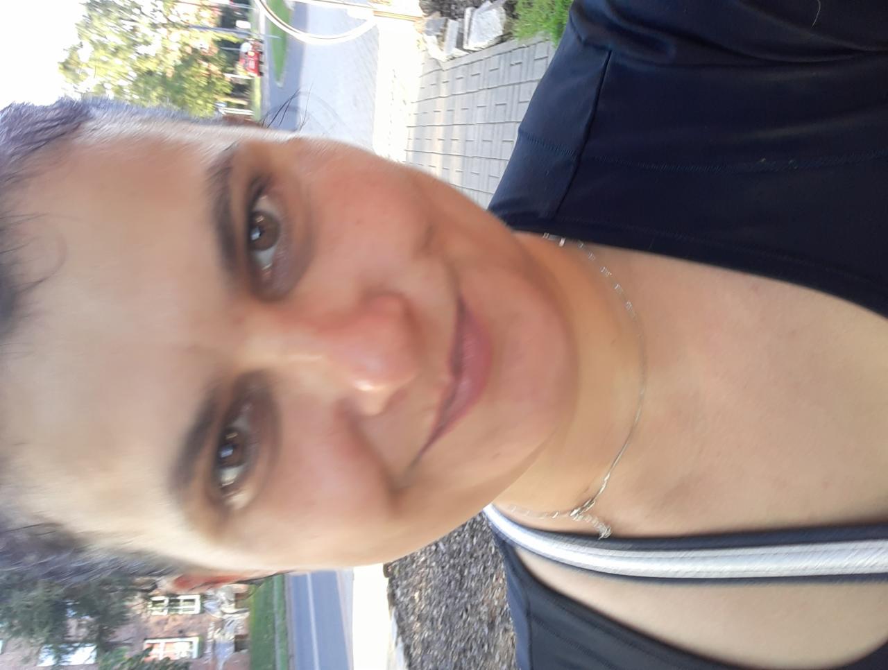 Uśmiechnięta kobieta w sportowym ubraniu, z delikatnym naszyjnikiem, sfotografowana z bliska. Widoczny fragment ulicy i zieleni w tle, zdjęcie obrócone o 90 stopni.