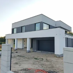 Nowoczesny dom z płaskim dachem w odcieniach szarości i bieli, z widoczną bramą garażową i elementami ogrodzenia w trakcie budowy, na tle błękitnego nieba.