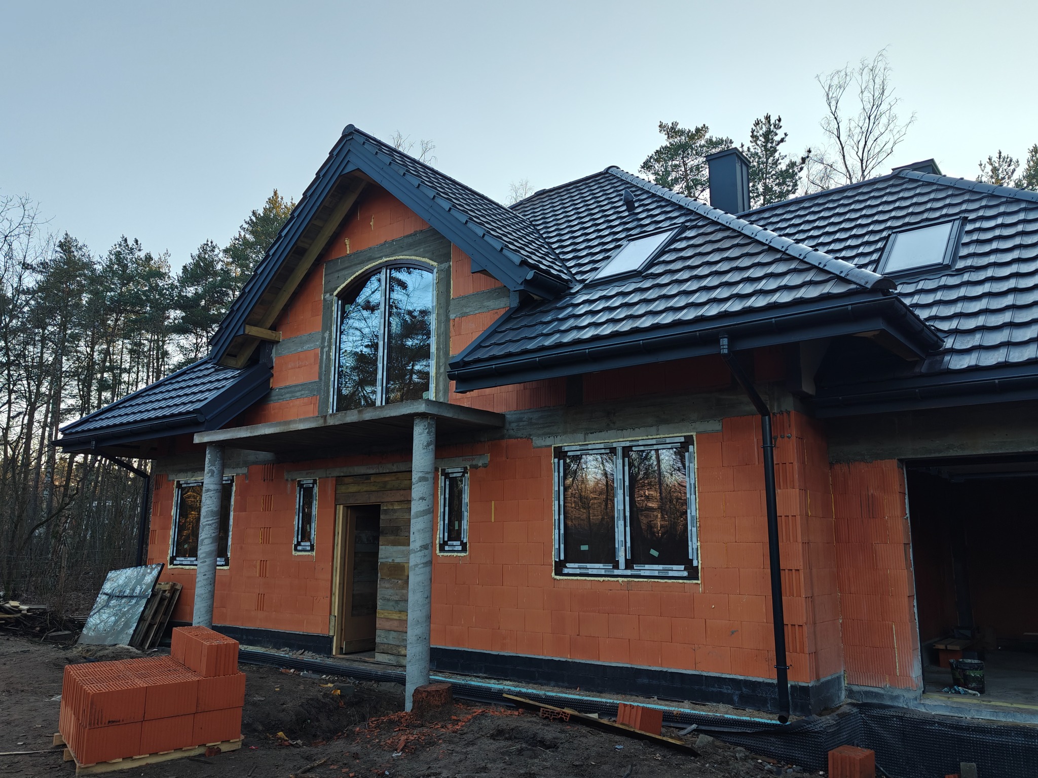 Dom w stanie surowym zamkniętym z czarną dachówką ceramiczną, widoczne okna dachowe i orynnowanie, na pierwszym planie paleta z pustakami.