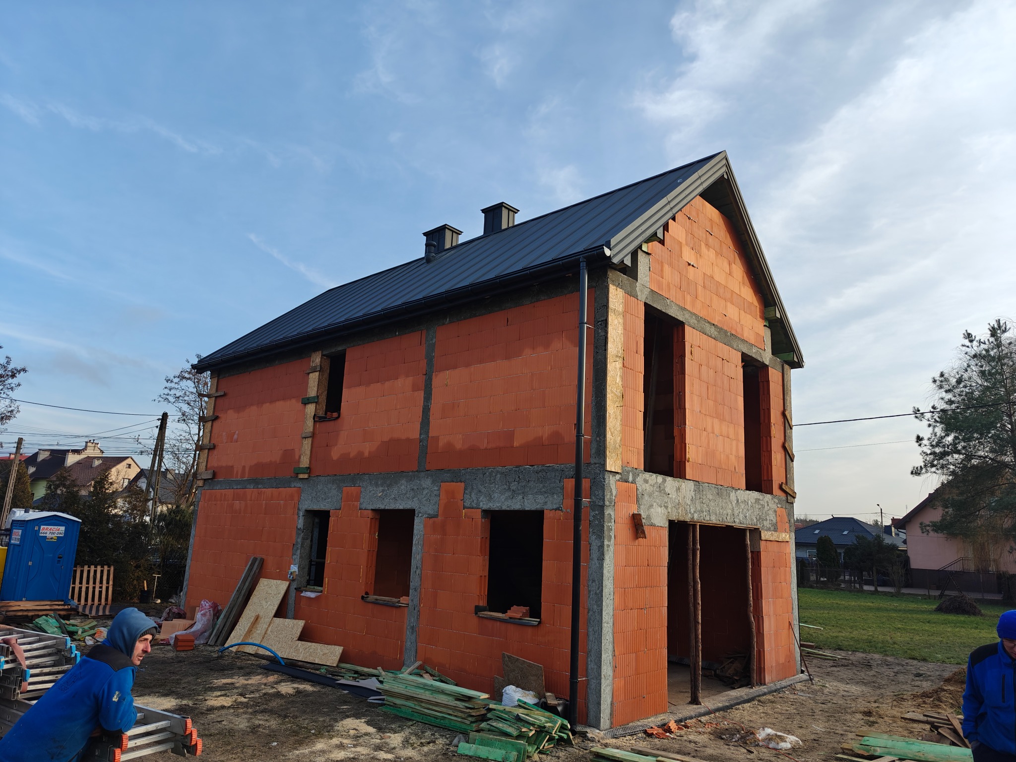 Surowy budynek w trakcie budowy z częściowo ukończonym dachem z blachy, widoczne bloczki ceramiczne i betonowe nadproża, pracownik opierający się o drabinę w lewym dolnym rogu.