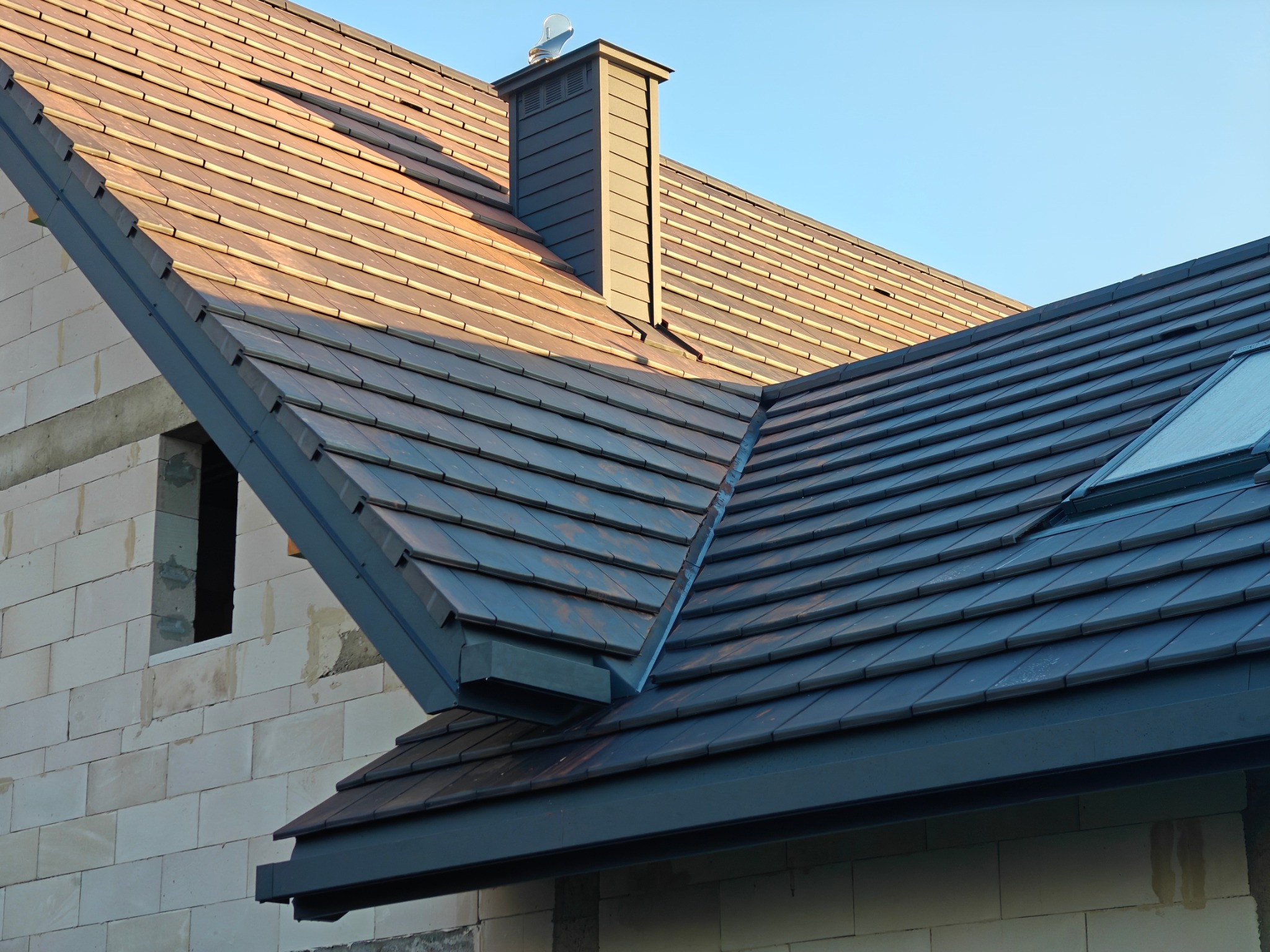 Nowe, grafitowe pokrycie dachowe z dachówki ceramicznej na budynku w trakcie budowy, widoczny komin i okno dachowe.