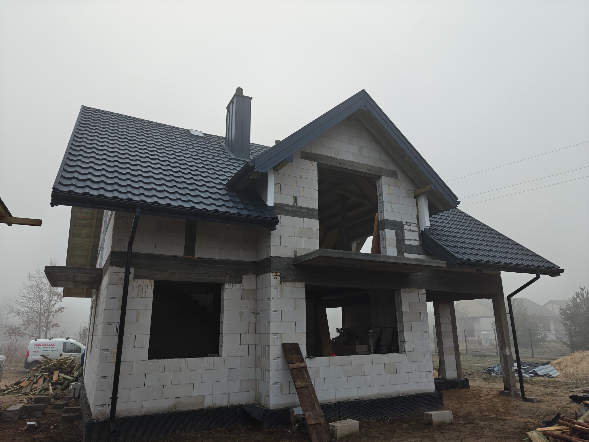 Dom w stanie surowym z czarnym dachem z blachy dachówkowej i kominem w mglisty dzień. Na pierwszym planie paleta i auto firmy dekarskiej.