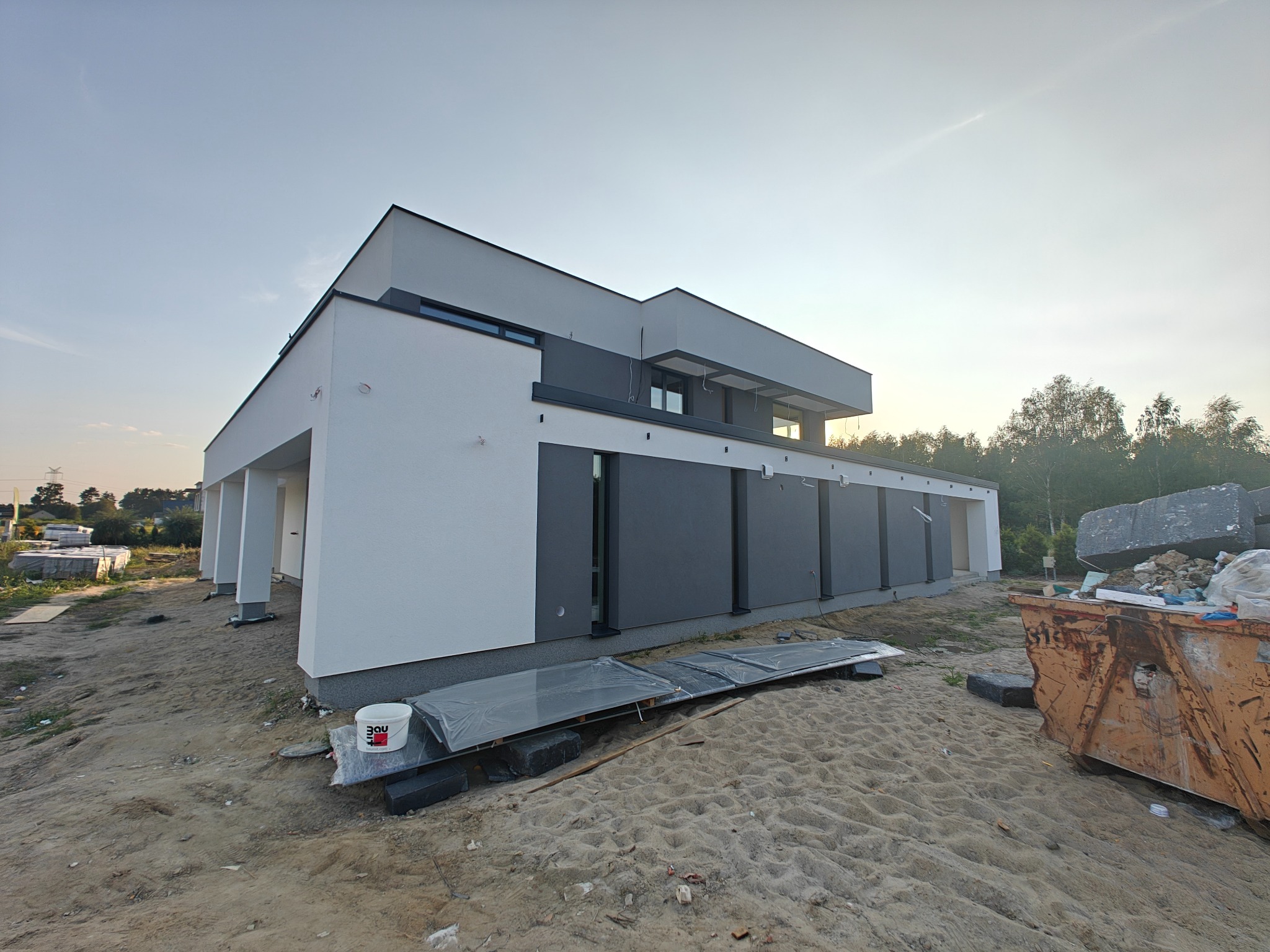 Nowoczesny dom jednorodzinny z płaskim dachem w trakcie budowy, widok z zewnątrz, z widocznymi materiałami budowlanymi i kontenerem na odpady na pierwszym planie.