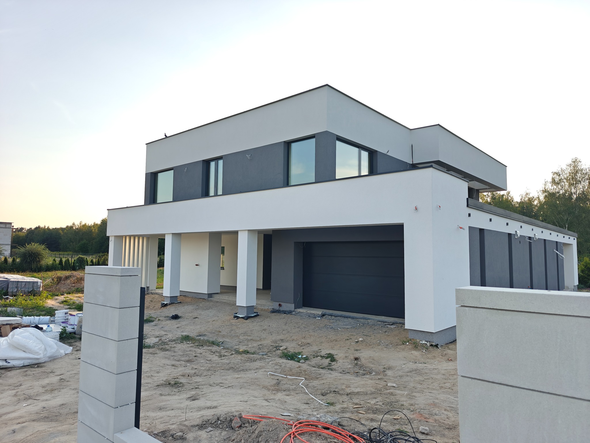Nowoczesny dom z płaskim dachem w odcieniach szarości i bieli, z widoczną bramą garażową i elementami ogrodzenia w trakcie budowy, na tle błękitnego nieba.