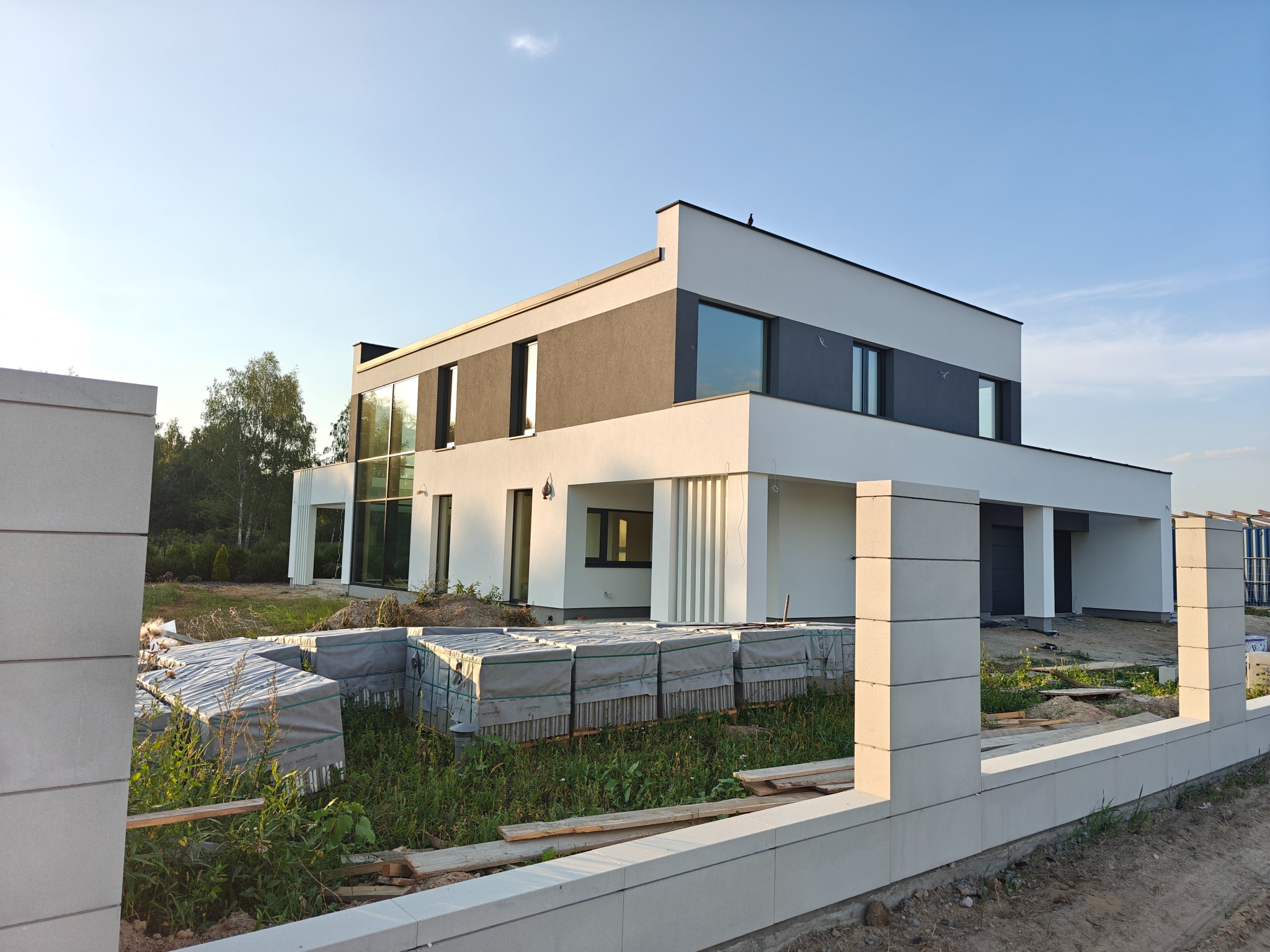 Nowoczesny dom z płaskim dachem w trakcie budowy, widoczne materiały budowlane i fragment ogrodzenia. Na dachu siedzi ptak. Ujęcie w słoneczny dzień.