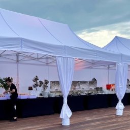 Catering Blue - Ustawione białe namioty cateringowe z bufetem i obsługą, widok z boku na drewnianym podeście, wieczorne oświetlenie.