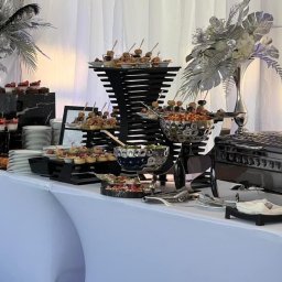 Catering Blue - Elegancki bufet z różnorodnymi przekąskami, deserami i daniami w metalicznych naczyniach, dekorowany srebrnymi kompozycjami kwiatowymi na białym tle draperii.