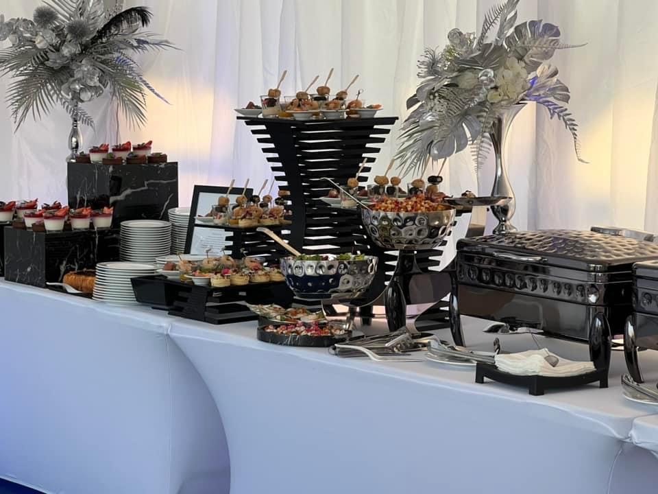 Elegancki bufet z różnorodnymi przekąskami, deserami i daniami w metalicznych naczyniach, dekorowany srebrnymi kompozycjami kwiatowymi na białym tle draperii.