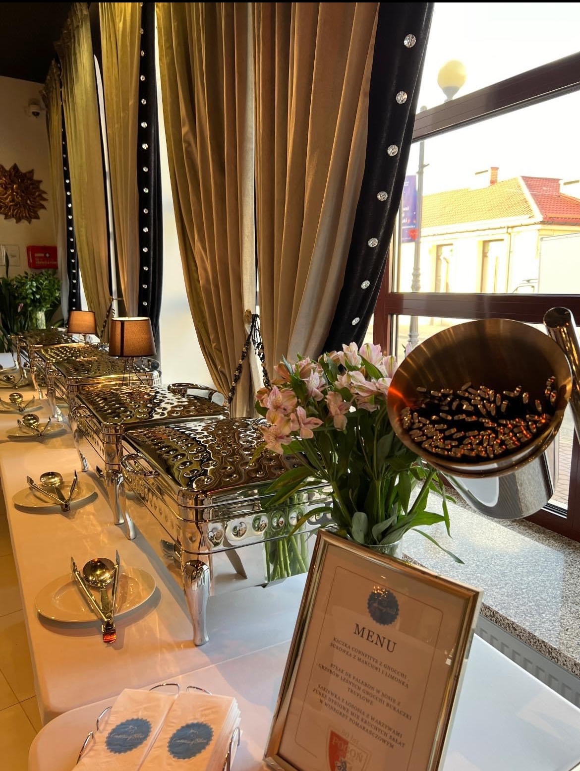 Elegancki bufet cateringowy z podgrzewaczami potraw, menu w ramce, kwiatami i dekoracyjnymi zasłonami w tle.