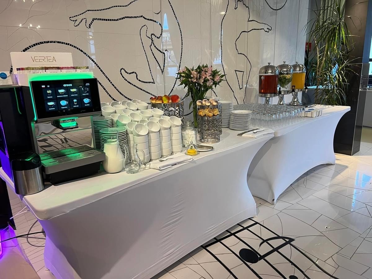 Stół cateringowy z białym obrusem: ekspres do kawy Vertea z menu na ekranie, stosy białych filiżanek, dzbanek z mlekiem, napoje w dystrybutorach, ciastka i kwiaty na tle ściany z mozaiką.