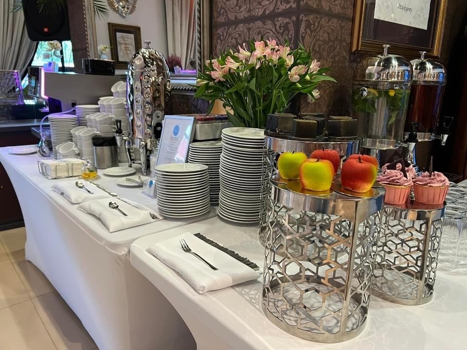Stół bufetowy z białym obrusem, zastawą stołową, napojami w dystrybutorach, jabłkami w cukrze i babeczkami, dekorowany kwiatami.