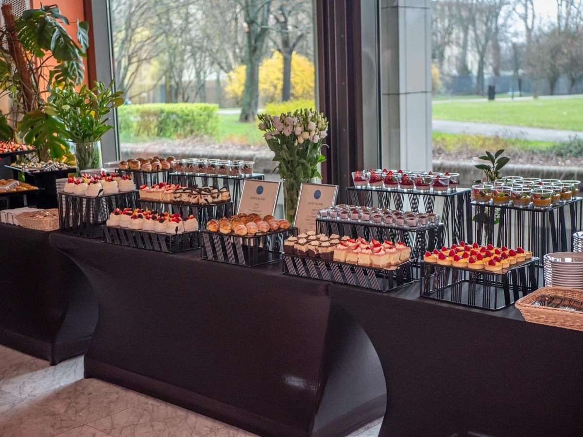 Stół z czarnym obrusem, na którym znajdują się metalowe stojaki z różnorodnymi deserami: mini tartaletki z malinami, ciasta z truskawkami, babeczki i desery w szklankach. W tle widoczne okno...