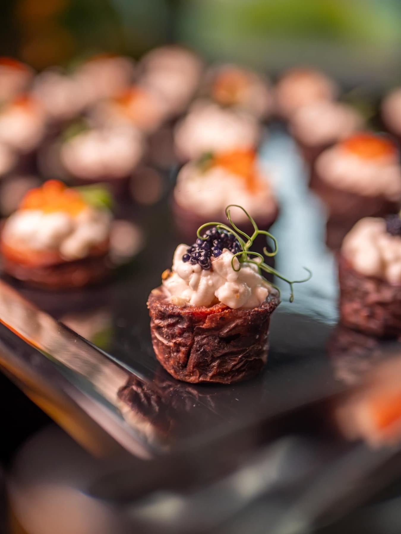 Miniaturowe tartaletki z kremowym nadzieniem i kawiorem na błyszczącej tacy, dekorowane zielonymi pędami.