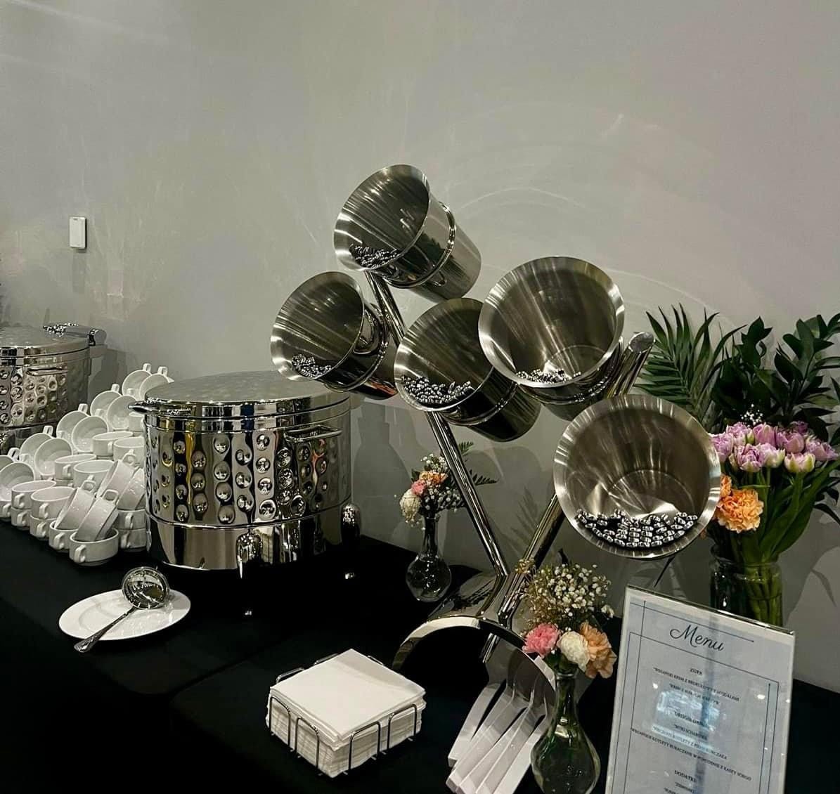 Elegancki stół cateringowy z błyszczącymi, metalowymi naczyniami na zupę, dekoracjami kwiatowymi i menu w ramce.