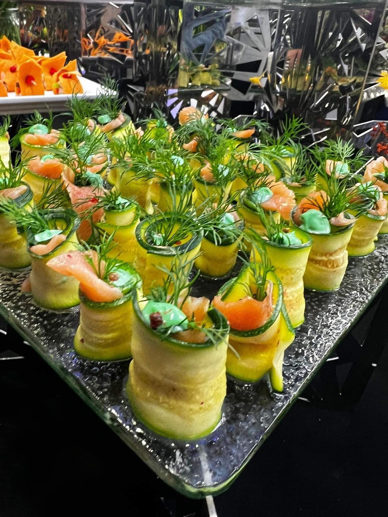 Elegancko ułożone roladki z cukinii z łososiem i koperkiem na szklanym talerzu, widoczne inne przekąski w tle.