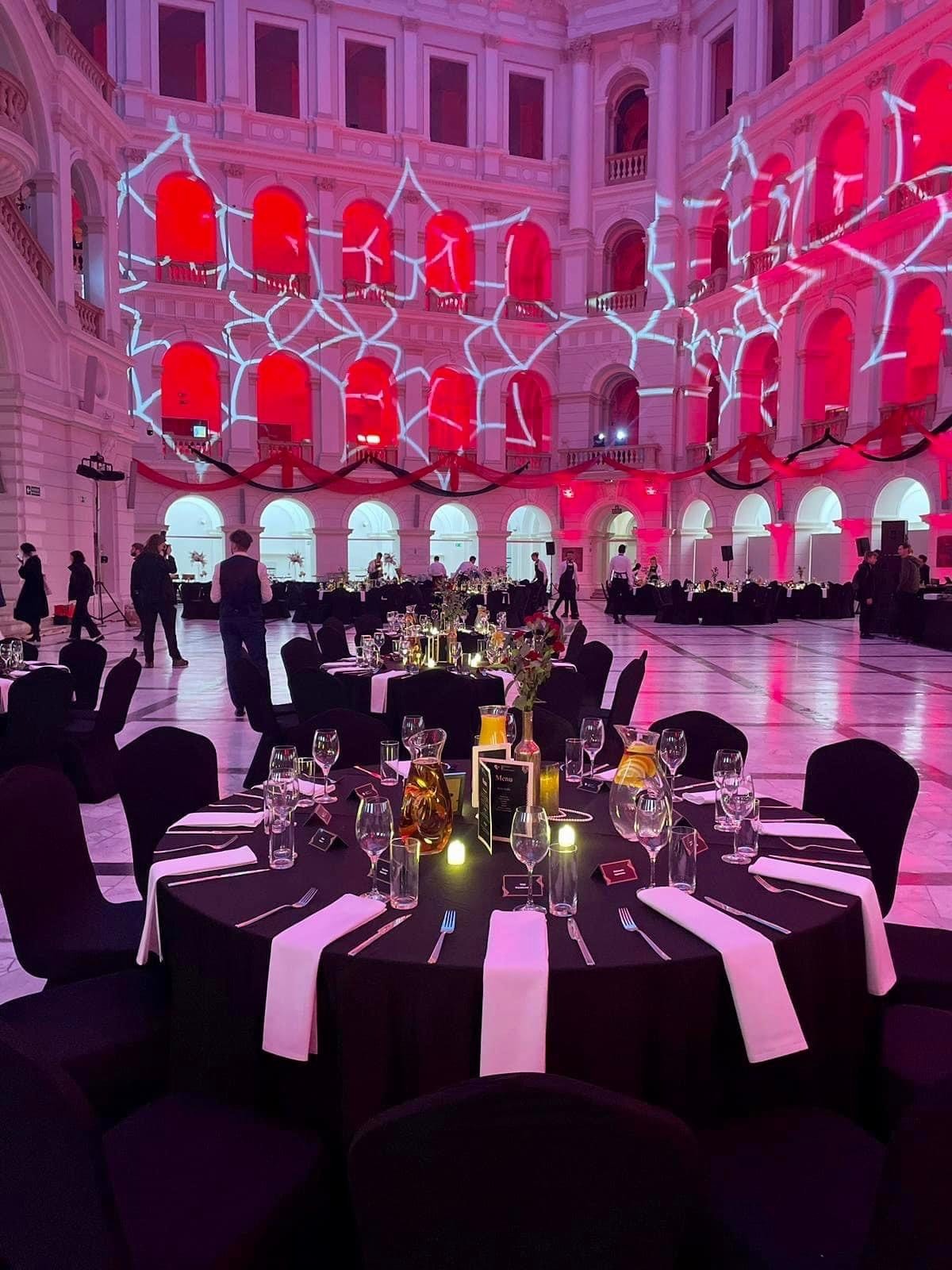 Elegancko nakryte okrągłe stoły z czarnymi obrusami i białymi serwetkami w dużej sali z czerwonymi iluminacjami na ścianach, przygotowane do uroczystości.