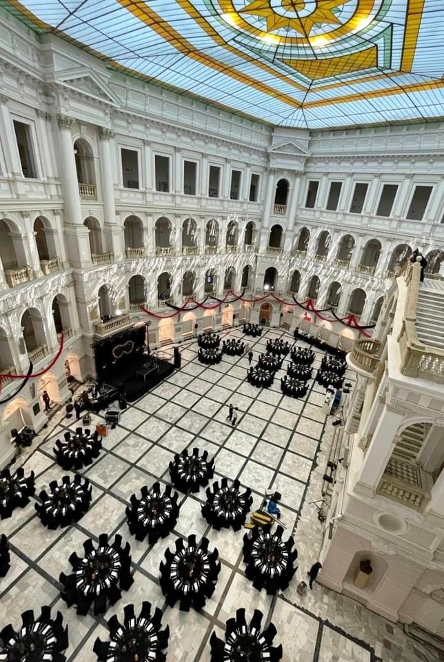 Przestronne wnętrze budynku z marmurową podłogą przygotowane na uroczystość, okrągłe stoły z czarnymi obrusami, scena z oświetleniem, widok z góry.