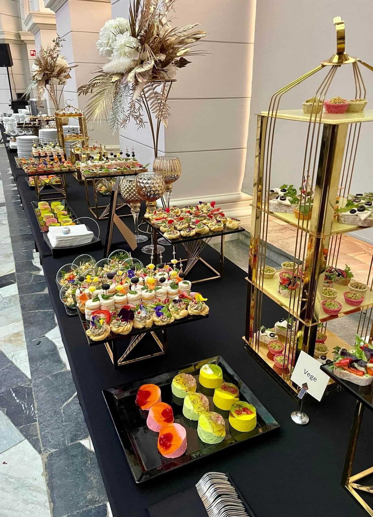 Elegancko zaaranżowany stół z różnorodnymi przekąskami, w tym tartaletki z kwiatami jadalnymi, mini kanapeczki i kolorowe desery, prezentowany w złotej klatce i na czarnych podkładkach,...