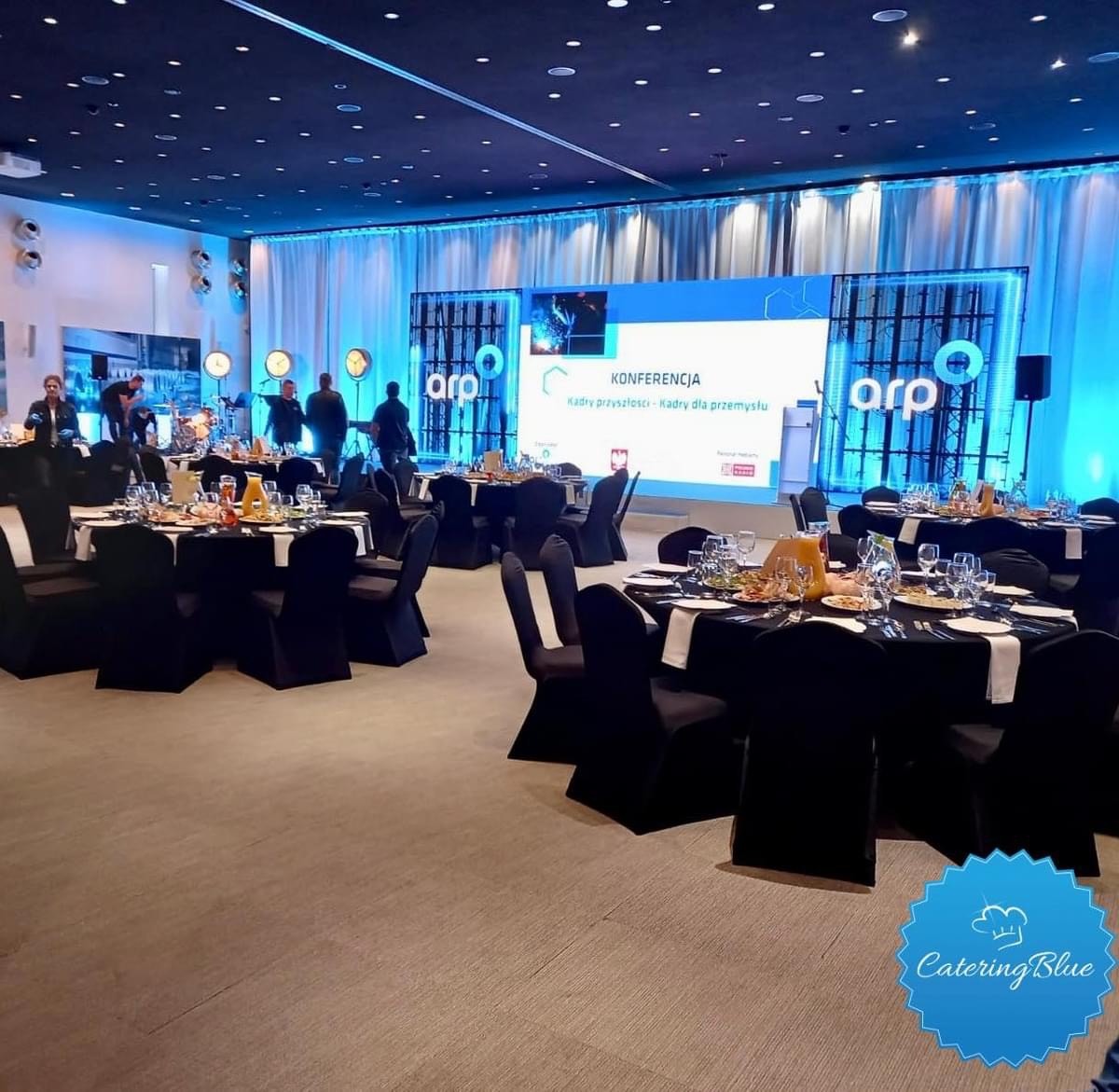 Sala konferencyjna z okrągłymi stołami nakrytymi czarnymi obrusami, białymi serwetkami i zastawą, przygotowana na przerwę kawową, z widocznym ekranem z logo ARP w tle.