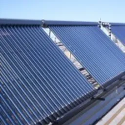 Rząd kolektorów słonecznych rurowych zamontowanych na dachu budynku, widocznych pod kątem, z jasnym niebem w tle.