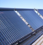 Rząd kolektorów słonecznych rurowych zamontowanych na dachu budynku, widocznych pod kątem, z jasnym niebem w tle.
