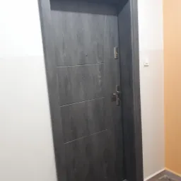 Szare drzwi wejściowe z wizjerem i trzema poziomymi frezami, zamontowane w ościeżnicy w korytarzu z pomalowanymi na biało i pomarańczowo ścianami.