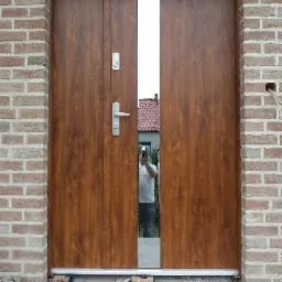 Brązowe drzwi wejściowe z wąskim przeszkleniem pośrodku, widoczna osoba robiąca zdjęcie w odbiciu, ceglana ściana w trakcie budowy.