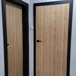 Dwa rodzaje drzwi wewnętrznych: pojedyncze otwierane i podwójne, z okleiną imitującą jasne drewno i czarnymi ościeżnicami oraz klamkami, na tle szarej ściany i podłogi.