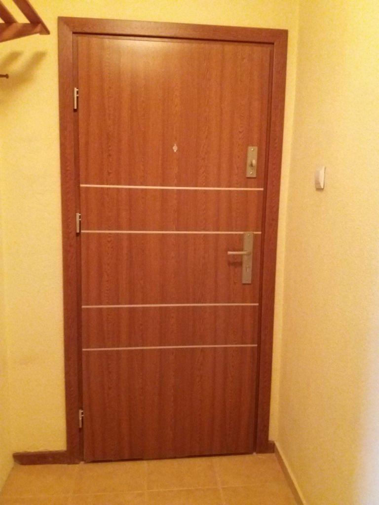 Brązowe drzwi wejściowe z czterema poziomymi, jasnymi intarsjami, wizjerem i klamką na tle żółtej ściany i podłogi z płytek.